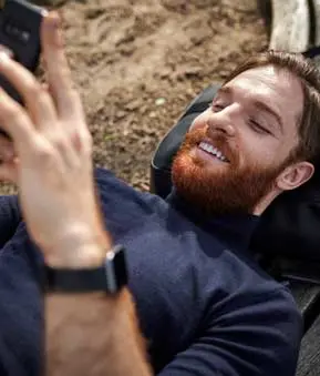 Person mit rötlich-braunem Bart liegt auf Holzbank, hält Smartphone in der rechten Hand und lächelt beim Blick auf den Bildschirm.