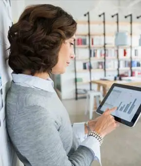 Person mit lockigen Haaren lehnt an Wand, hält Tablet in der Hand und blickt auf ein Balkendiagramm.
