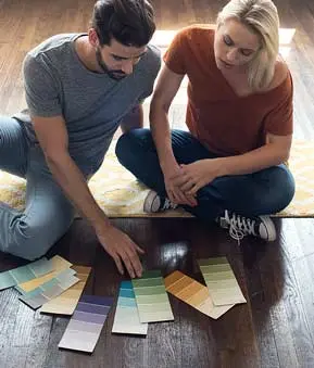 Zwei Personen sitzen auf dem Boden einer Wohnung, betrachten gemeinsam einen Farbfaecher. Im Hintergrund helle Wände und Renovierungsutensilien.