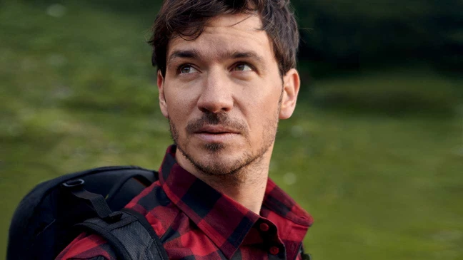 Felix Neureuther in kariertem Hemd kniet am Seeufer und hält die Hände ins Wasser, im Hintergrund bewaldete Hügel.