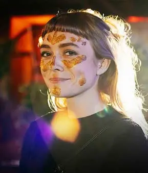 Person mit goldenem Gesichtsmake-up und schwarzem Kleidungsstück blickt in die Kamera. Hintergrund ist unscharf.