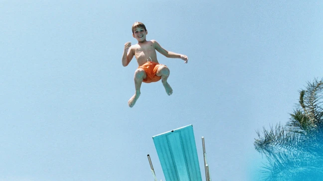 Junge springt von einem Sprungbrett in einen Pool. Im Hintergrund blauer Himmel und eine Palme.