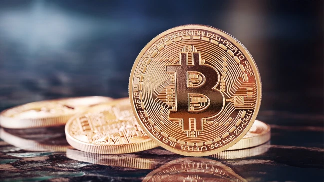 Nahaufnahme einer goldfarbenen Bitcoin‑Münze mit dem typischen ‚B‘‑Symbol, die auf weiteren gestapelten Münzen liegt; glänzende Metalloberfläche auf reflektierendem Untergrund.