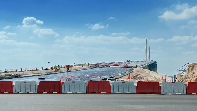 Baustelle einer großen Straßen- oder Brückenkonstruktion mit breiten Fahrspuren, rot‑weißen Absperrbarrieren und mehreren Arbeiterinnen und Arbeitern im Hintergrund unter blauem Himmel.