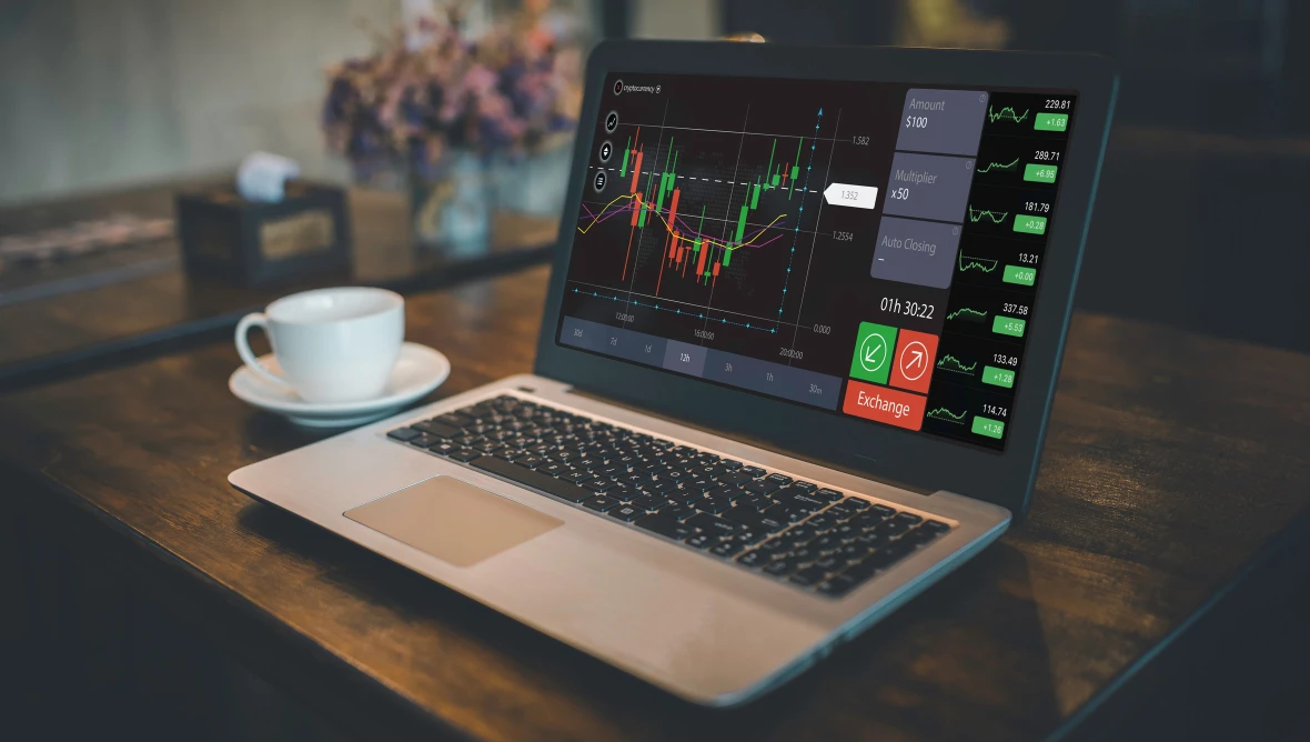 Offenes Laptop auf einem Holztisch mit einer Handels‑ und Kurschart‑Software auf dem Bildschirm; daneben stehen eine Tasse Kaffee und eine Vase mit Blumen