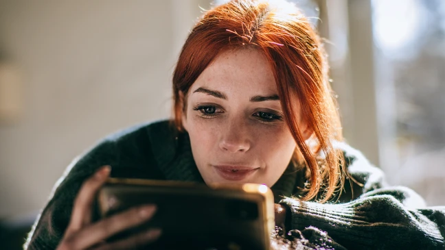 Person mit langen Haaren sitzt in Innenraum vor hellem Fenster und hält ein Tablet mit beiden Händen in Vorderansicht gezeigt.