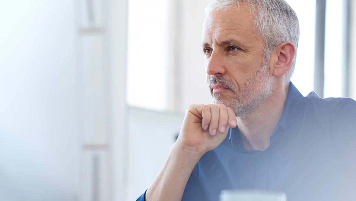 Porträt einer nachdenklichen älteren Person mit grauem Haar und Bart im hellen Raum.