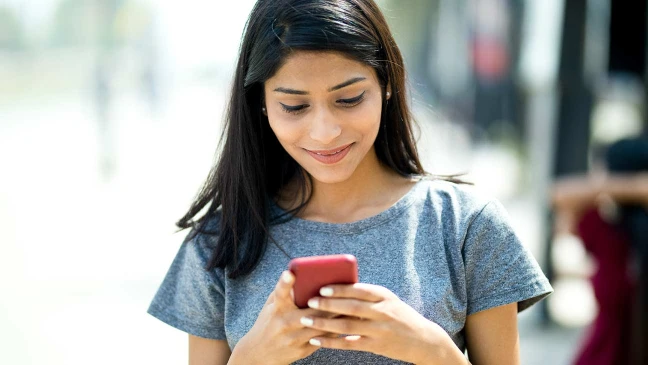 Person mit langen Haaren in grauem T-Shirt blickt draußen lächelnd auf ein rotes Smartphone in den Händen.