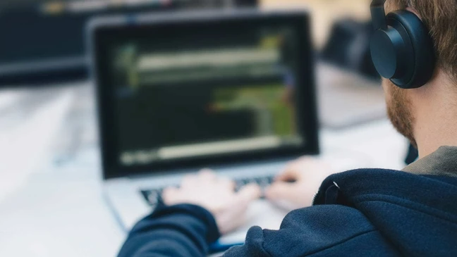 Person mit Kopfhörern sitzt vor einem Laptop, der Textzeilen zeigt, in einem unscharfen Hintergrund.