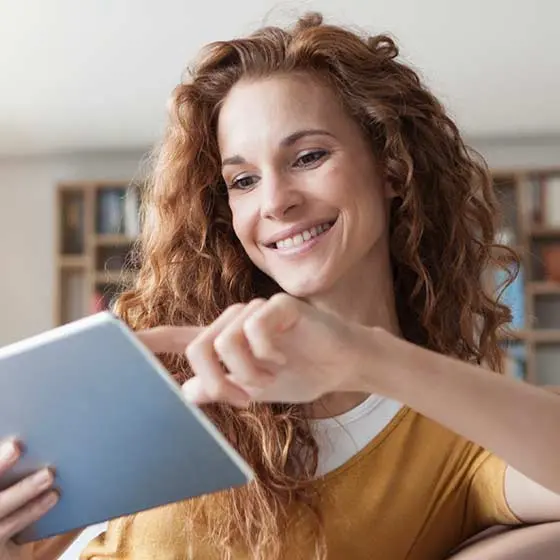 Lachende Person mit lockigem Haar bedient ein Tablet in einem Raum mit Bücherregalen im Hintergrund.