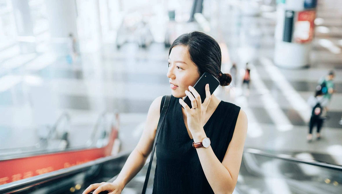 Person mit dunklem Haar steht auf einer Rolltreppe in einem modernen Gebäude und telefoniert mit einem Smartphone.
