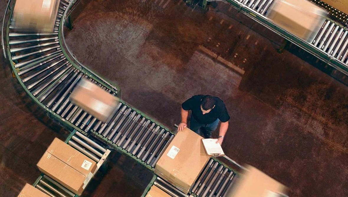 Blick von oben auf eine Person an einem Förderband, die ein Paket prüft. Mehrere Kartons bewegen sich auf dem Band.